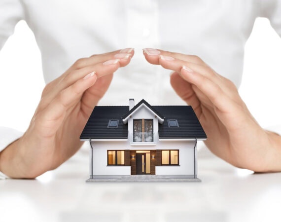 A person gently cups their hands protectively over a small model house, symbolizing home security, protection, or insurance—an image often associated with Elder Law in Smithtown, NY. The background is plain and the focus is on the hands and the house.