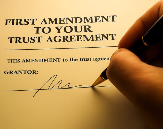 A hand holding a pen signs a document titled "First Amendment to Your Trust Agreement," ready for the grantor’s signature. Warm lighting highlights this moment, perfect for Elder Law guidance in Smithtown, NY.